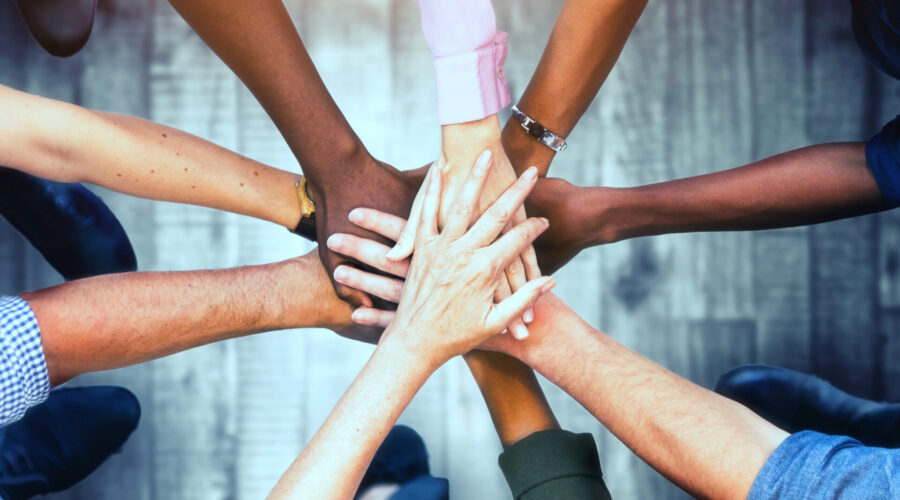 Overhead view of arms and hands reaching to a central point, like spokes on a wheel. The hands, representing various races or ethic groups, are all laid one on top of the other.